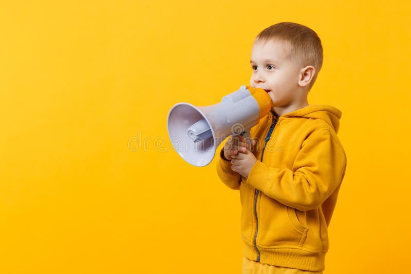 Little fun kid boy 3-4 years old in yellow clothes holding, speaking in electronic megaphone isolated on orange wall