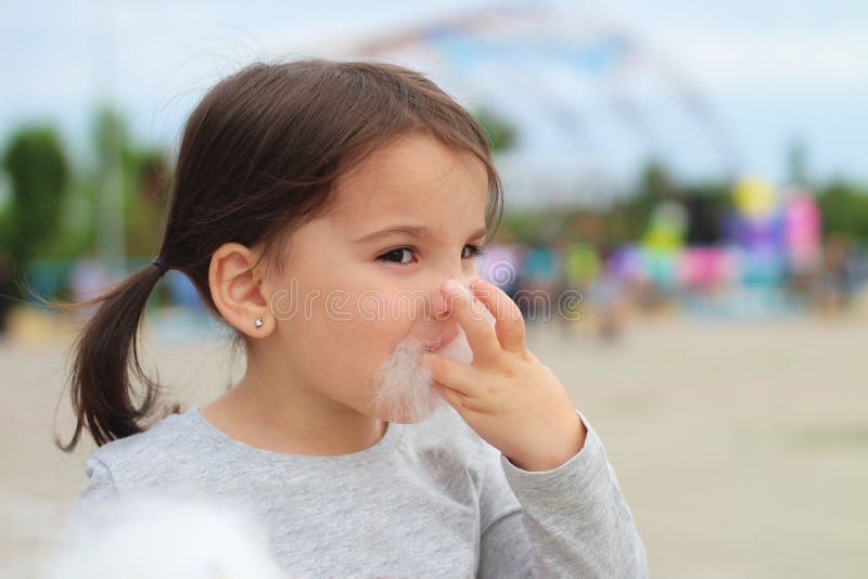 Little funny white girl with tails with narrow eyes holds cotton candy with his lips while walking outdoors stock image Little funny white girl with tails with narrow eyes holds cotton candy with his lips while walking outdoors stock image