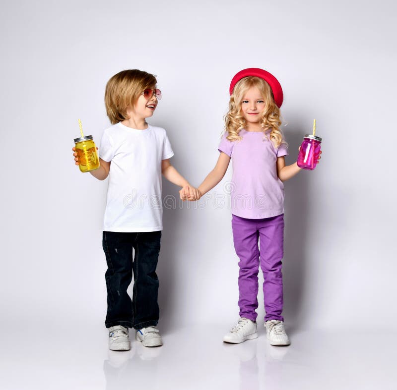 Little girl and boy in colorful casual clothes. Smiling, holding hands, yellow and pink cocktail bottles. Posing isolated on white royalty free stock photography Little girl and boy in colorful casual clothes. Smiling, holding hands, yellow and pink cocktail bottles. Posing isolated on white royalty free stock photography
