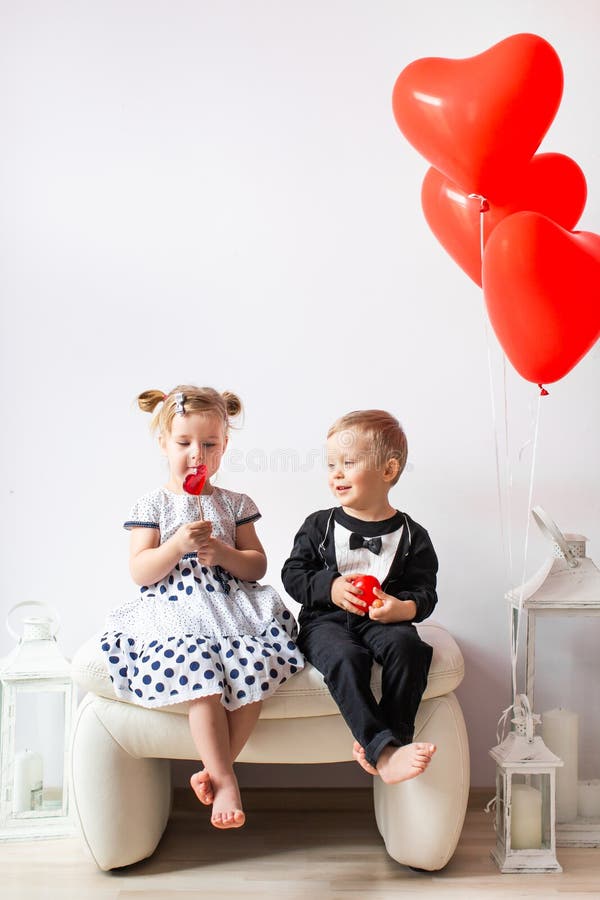 Little girl and boy sitting on a white chair near heart-shaped baloons. Girl licking a red lollipop. Valentines day