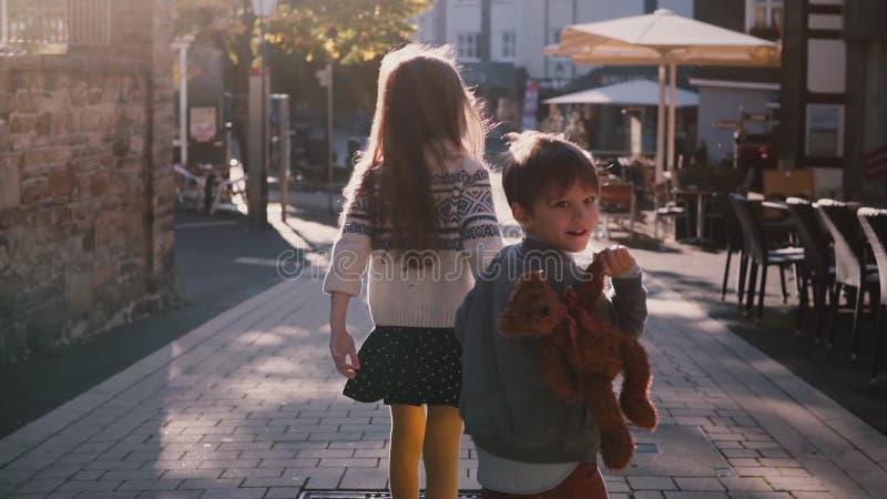 Little girl and boy walk together holding hands. Slow motion. Back view. Two kids wander around old town on a sunny day.