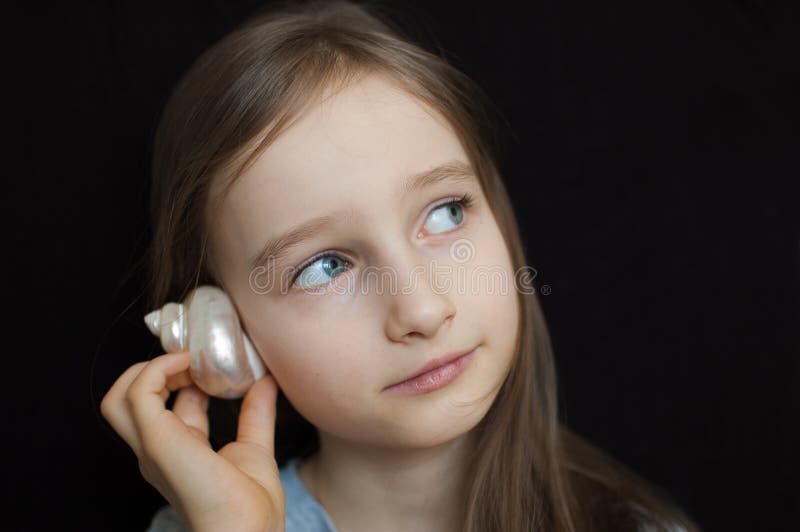 Little girl is dreaming about the summer trip to the ocean and listening to a white shell to hear a sea sound on black