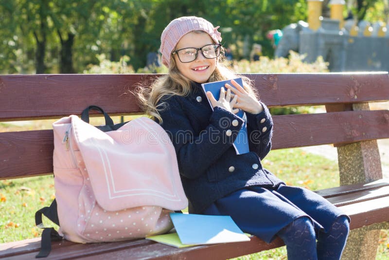 Little girl elementary school student sitting on bench with backpack, looks at school books and notebooks. Background autumn city park.