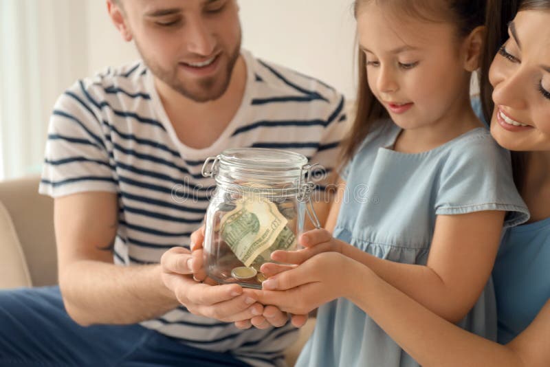 Little girl with her parents holding glass jar with dollars indoors. Money savings concept