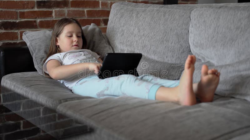 A Little Girl Lies at Home on the Couch and Looks into the Black Tablet. the Child Plays and Watches the Video Stock Video - Video of person, laptop: 215211925