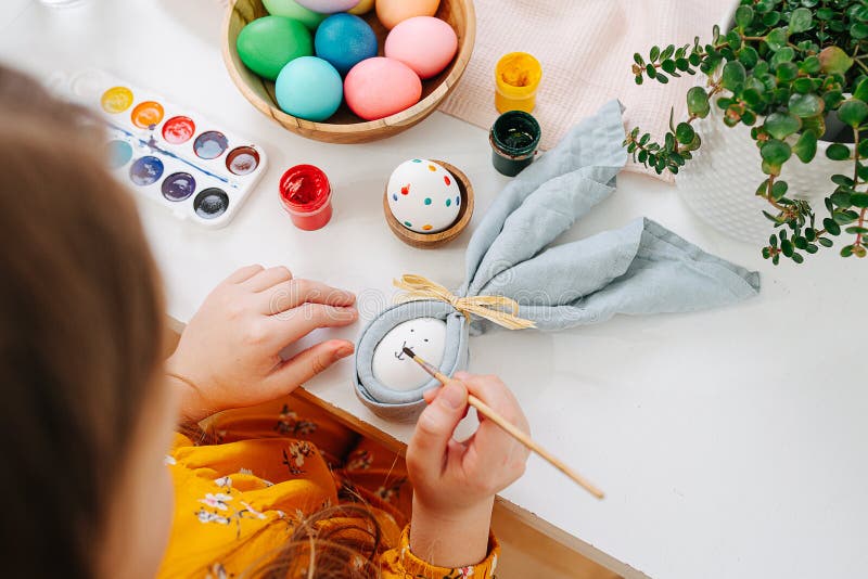 Little girl painting easter eggs behind a table using brush. Top view.