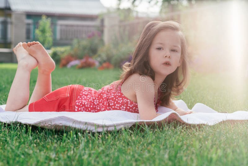 Little 3-4 girl in red clothes lies on blanket on green grass looking into frame