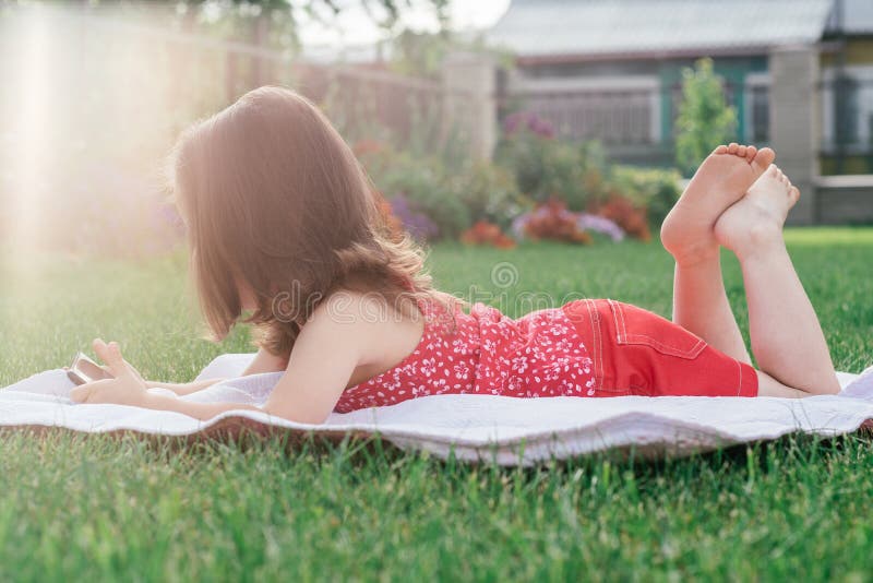 Little 3-4 girl in red clothes lies on blanket on green grass and looks into mobile phone. Children, using gadgets