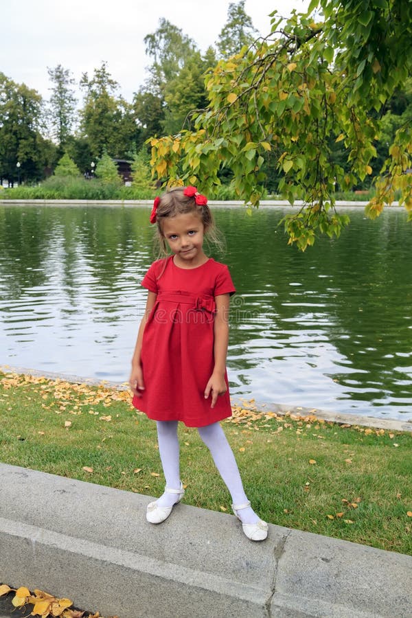 little girl with red hairpins and dress stands near pond outdoor in autumn