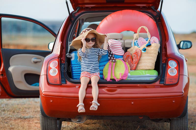 Little girl , brunette with long curly hair , dressed in a striped sailor's shirt , dark sunglasses and a large straw hat,goes on a journey to the sea , sits in the trunk of the red car loaded with suitcases and bags. Little girl , brunette with long curly hair , dressed in a striped sailor's shirt , dark sunglasses and a large straw hat,goes on a journey to the sea , sits in the trunk of the red car loaded with suitcases and bags