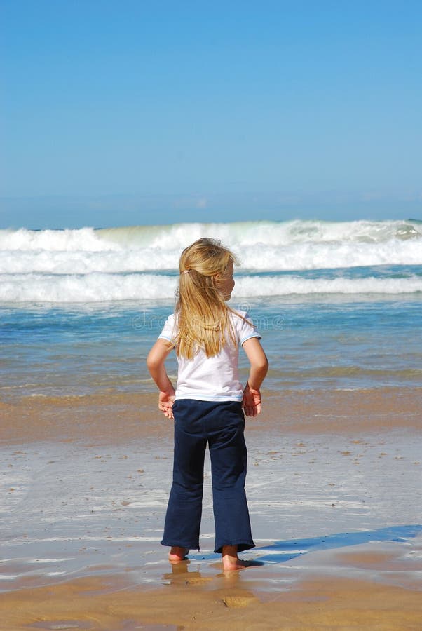 Rear view of a cute little caucasian girl child standing barefoot in the wet sand of a beach and watching the wild blue sea. Rear view of a cute little caucasian girl child standing barefoot in the wet sand of a beach and watching the wild blue sea