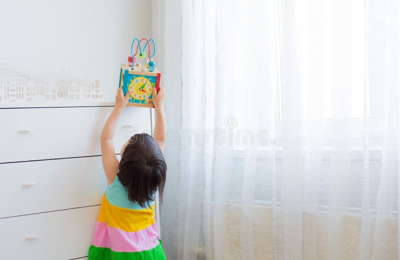 A little girl 3 years stretches to get an educational toy from a high shelf. Bright dress on the white background of the room. Free space. Out of reach of children, child safety