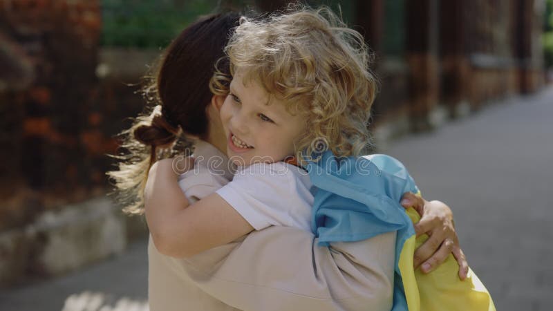 Little happy ukrainian child with ukrainian flag on shoulders hugs his mother on the street. near school. Stop war in