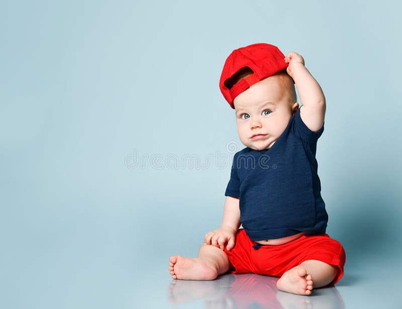 Little kid in t-shirt, red shorts and cap, barefoot. He holding a rattle, sitting on floor against blue background. Close up