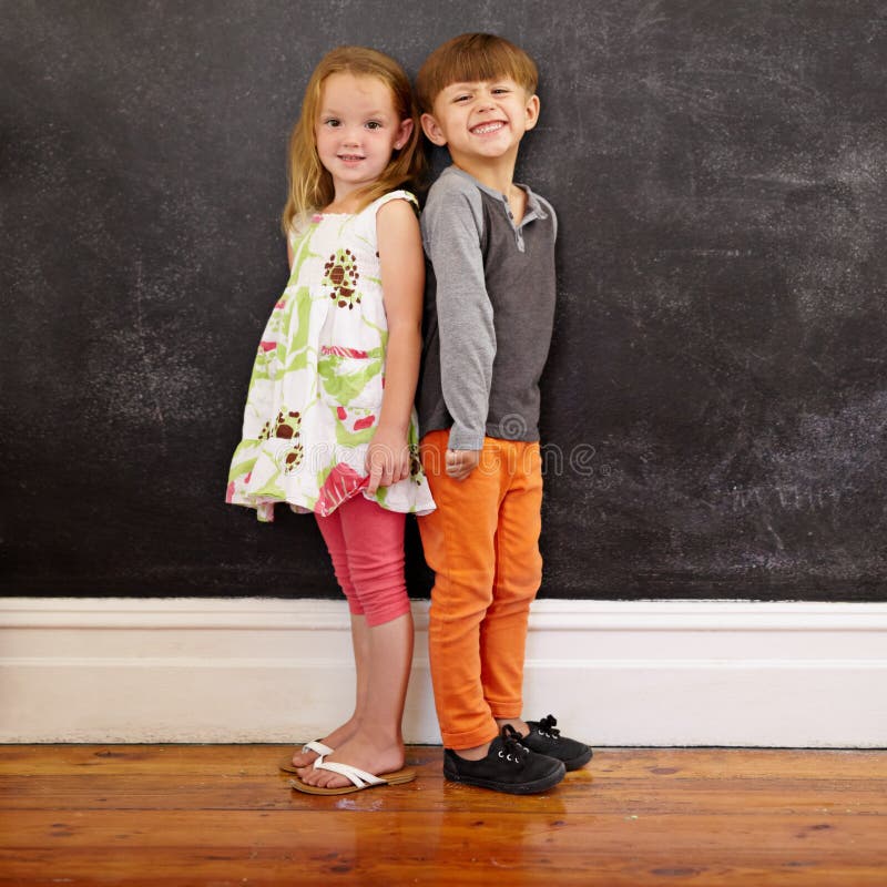 Little boy and girl standing back to back in front of blackboard looking at camera smiling. Full length image of two innocent little children together at home. Little boy and girl standing back to back in front of blackboard looking at camera smiling. Full length image of two innocent little children together at home.