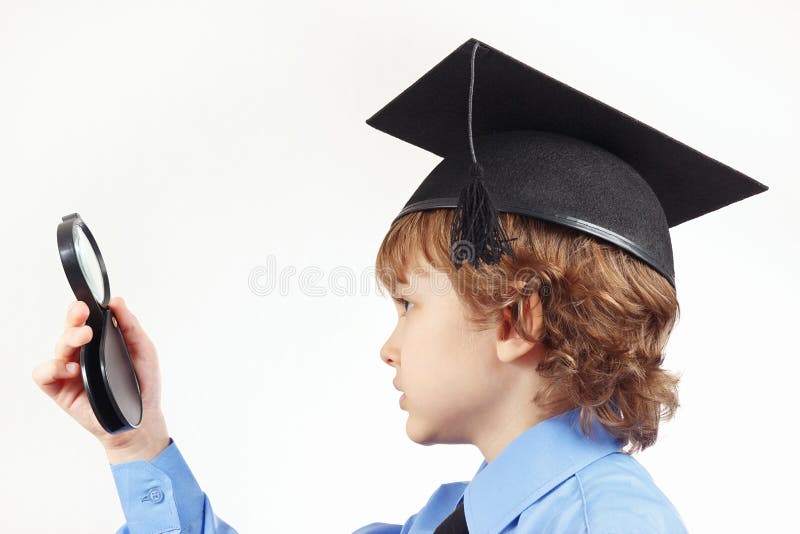 Little pensive boy in academic hat with a magnifying glass on white background.