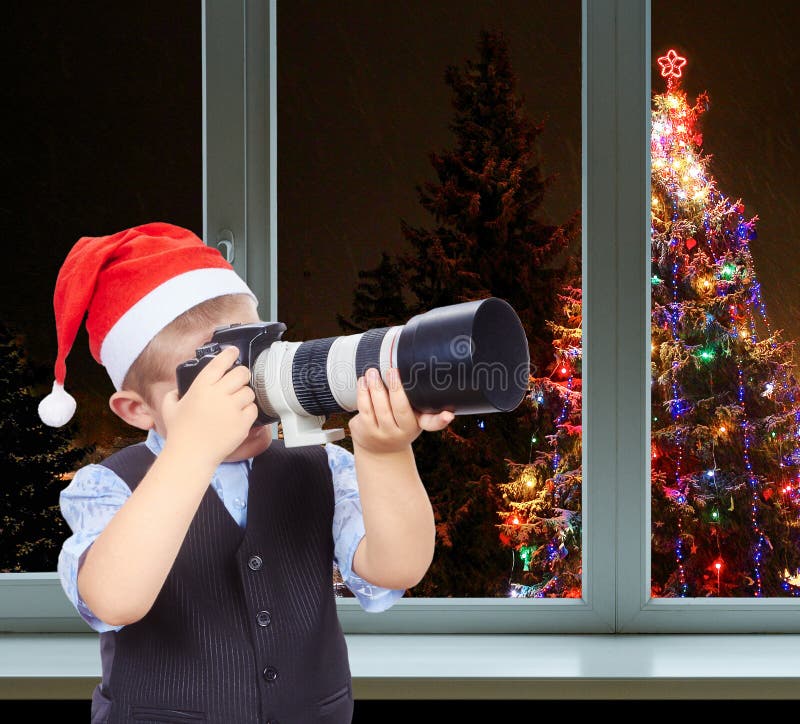 Little photographer in cap of Santa Claus on a background decorated Christmas tree outside the window royalty free stock photo Little photographer in cap of Santa Claus on a background decorated Christmas tree outside the window royalty free stock photo