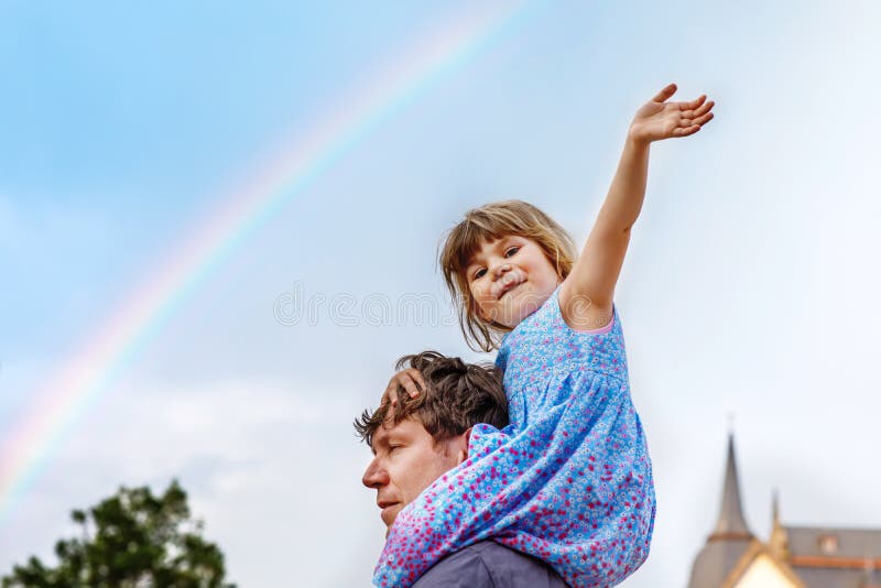 Little preschool girl sitting on shoulder of father. Happy toddler child and man observing rainbow on sky after summer