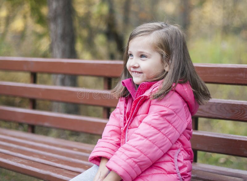 Little pretty girl in a pink coat sitting on a wood bench at the park in autumn. Emotional portrait royalty free stock image Little pretty girl in a pink coat sitting on a wood bench at the park in autumn. Emotional portrait royalty free stock image