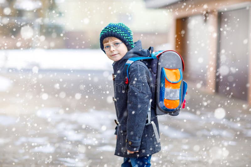 Little school kid boy of elementary class walking to school. Happy child having fun and playing with first snow. Student with backpack in colorful winter clothes. School building