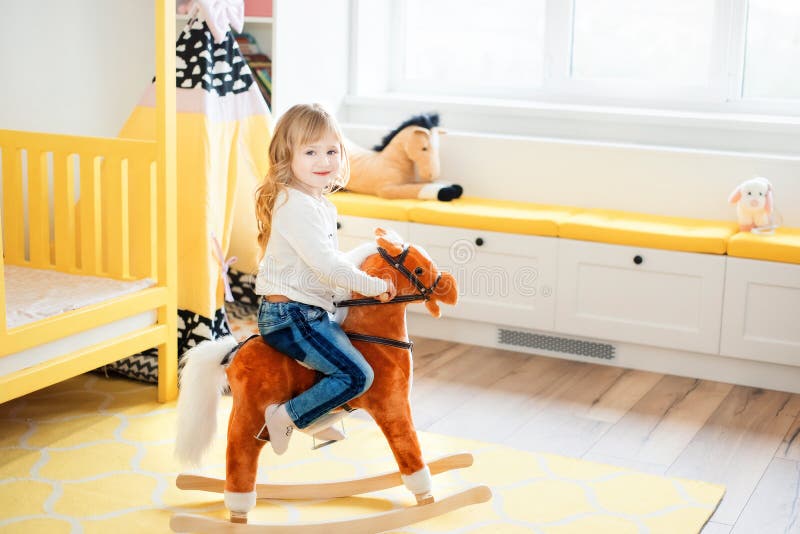 Little smiling blonde 3 years old girl sitting on rocking horse in yellow bedroom at home in day time. Time to play during self