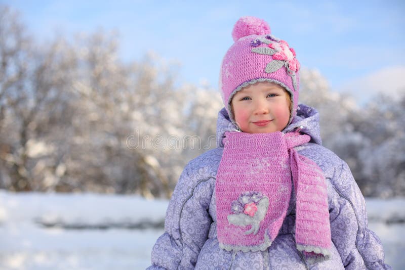 Little smiling girl in pink scarf and hat stands near trees