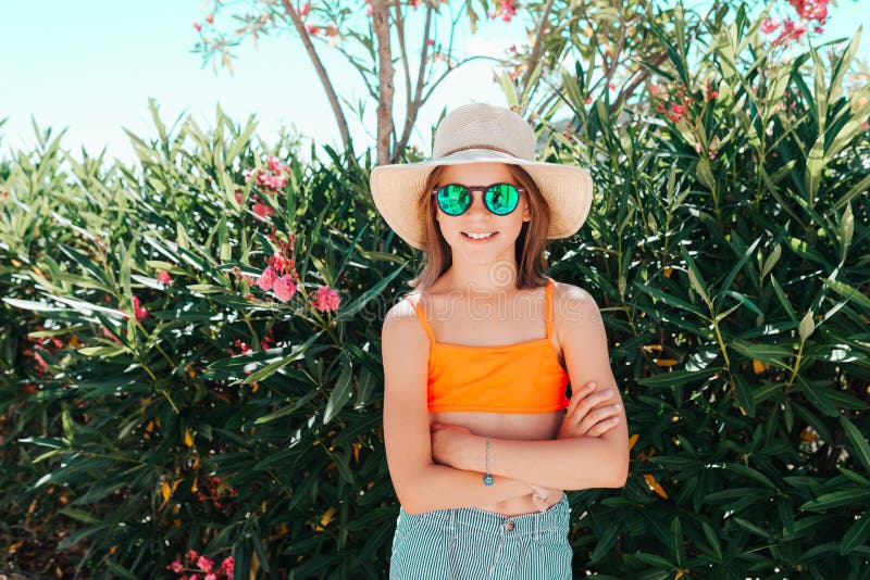 Little smiling playful girl in a straw hat and sunglasses crossing her arms in front of her - looking directly at the camera -