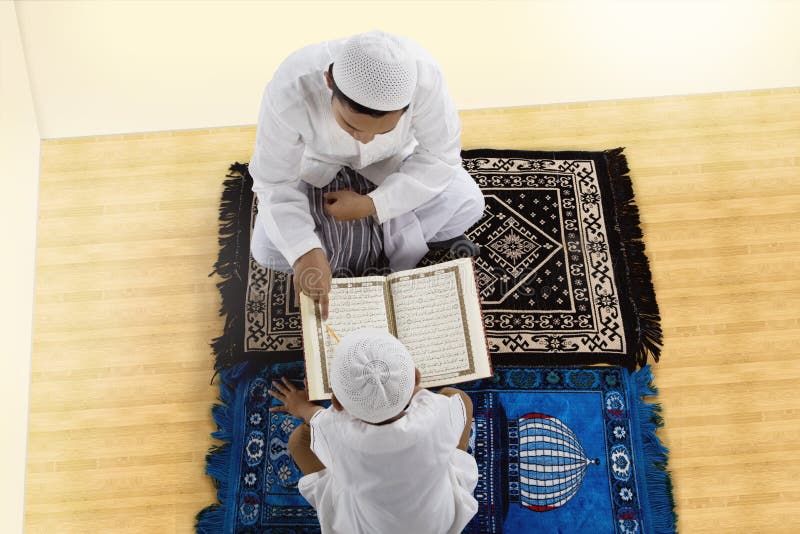 High angle of little son and father reading Quran together while sitting on prayer carpet at home