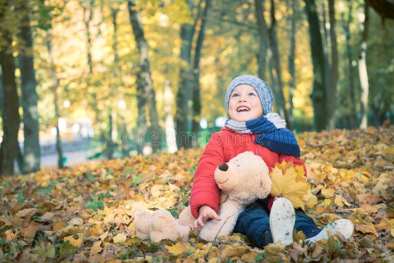 Little toddler boy, sitting with teddy bear in the autumn park, looking at throwing leaves around himself.