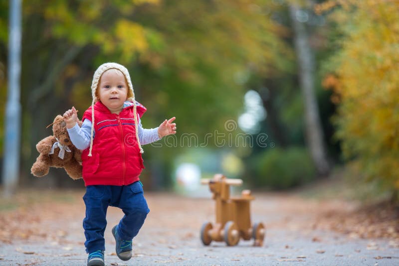 Little toddler boy with teddy bear, riding wooden dog balance bike in autumn park on a sunny warm day, children leisure activities and happiness concept