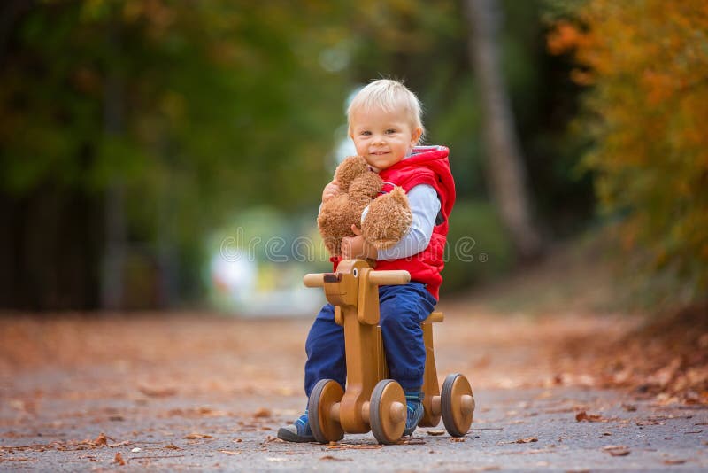 Little toddler boy with teddy bear, riding wooden dog balance bike in autumn park on a sunny warm day, children leisure activities and happiness concept