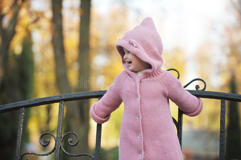 Little cute 3-4-year-old blonde girl in a knitted coat with a hood stands on a bench and smiles against the backdrop of