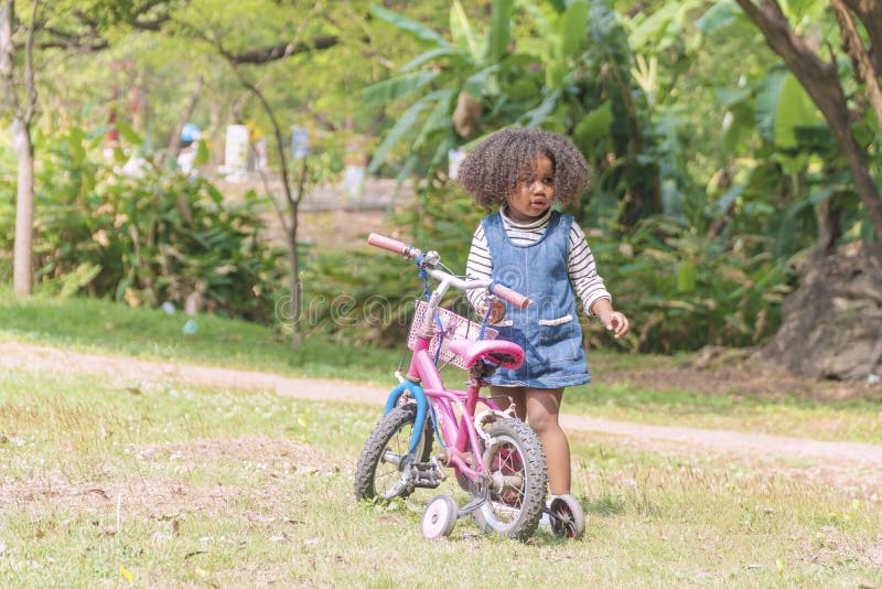 Black girl want learn ride bicycle while standing on the green grass in the park. Mixed race kid girl hold