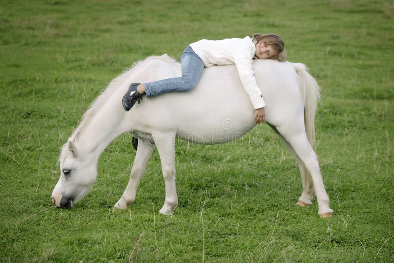 Little young girl in a white sweater and jeans lying backwards on the back of a white horse. Lifestyle portrait