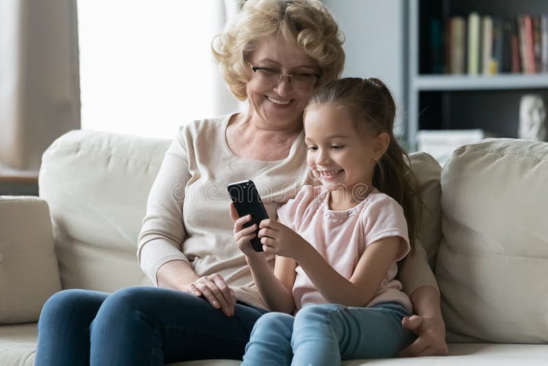 Little girl holding cellphone sitting with grandmother on sofa. In living room sit on sofa resting little granddaughter with old grandma, kid holds cell phone stock images
