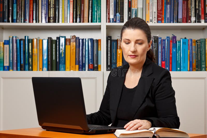 Mature woman in an office or library with her laptop reading in a book, advocate, lawyer, teacher, prof, historian, psychologist, diplomat, translator. Mature woman in an office or library with her laptop reading in a book, advocate, lawyer, teacher, prof, historian, psychologist, diplomat, translator