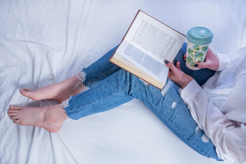Livro da terra arrendada da estudante nos pés com pés desencapados e chá quente de leitura e bebendo na cama acolhedor