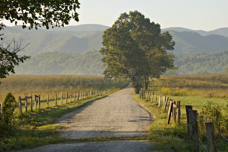 Beautiful morning light on gravel road in Great Smoky Mountains National Park. Road is lined by natural wood fence and beautiful trees. Mountains and fog are in the background. Beautiful morning light on gravel road in Great Smoky Mountains National Park. Road is lined by natural wood fence and beautiful trees. Mountains and fog are in the background.