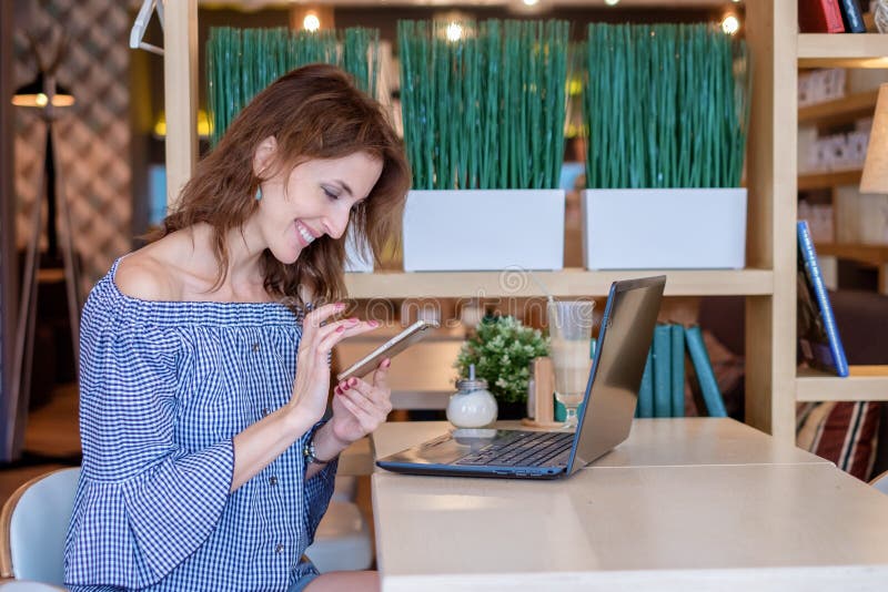 Lle free lance femminili stanno parlando su un telefono mentre si sedevano in un caffè con un computer portatile su una tavola Gi fotografie stock libere da diritti Lle free lance femminili stanno parlando su un telefono mentre si sedevano in un caffè con un computer portatile su una tavola Gi fotografie stock libere da diritti