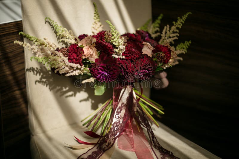 Lle nozze hanno arruffato il mazzo con le dalie di Borgogna e le rose color crema con i nastri sta su una sedia, accesa dal sole immagine stock Lle nozze hanno arruffato il mazzo con le dalie di Borgogna e le rose color crema con i nastri sta su una sedia, accesa dal sole immagine stock