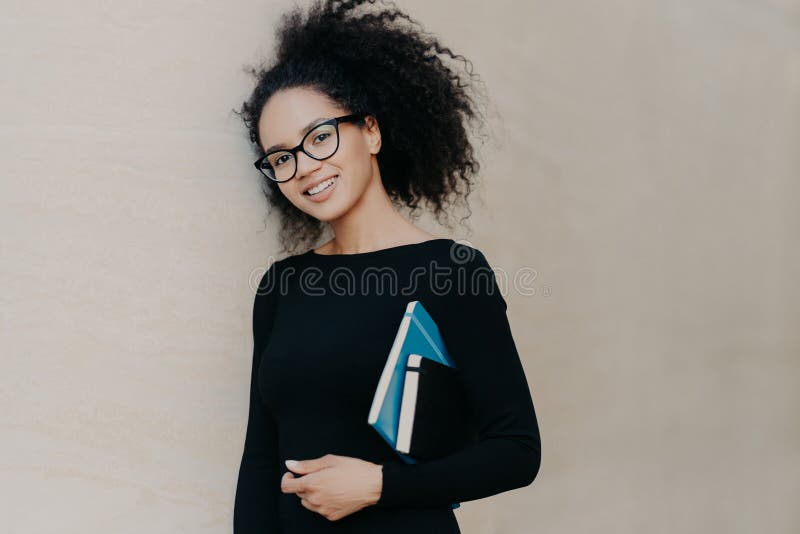 Lo studente afroamericano riccio positivo porta il blocco note ed il diario, indossa i vestiti neri, vetri trasparenti, gode di d immagine stock Lo studente afroamericano riccio positivo porta il blocco note ed il diario, indossa i vestiti neri, vetri trasparenti, gode di d immagine stock