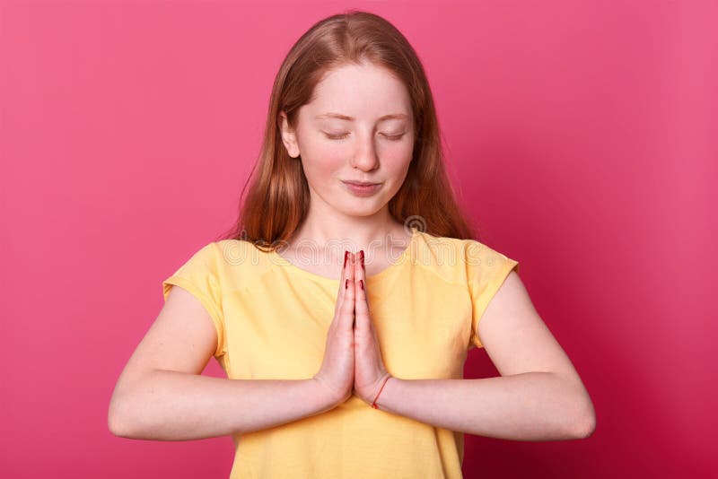 Lo studio sparato di bella ragazza caucasica calma dell'adolescente sta isolato sopra fondo rosa mentre prega, tiene gli occhi ch immagini stock libere da diritti Lo studio sparato di bella ragazza caucasica calma dell'adolescente sta isolato sopra fondo rosa mentre prega, tiene gli occhi ch immagini stock libere da diritti