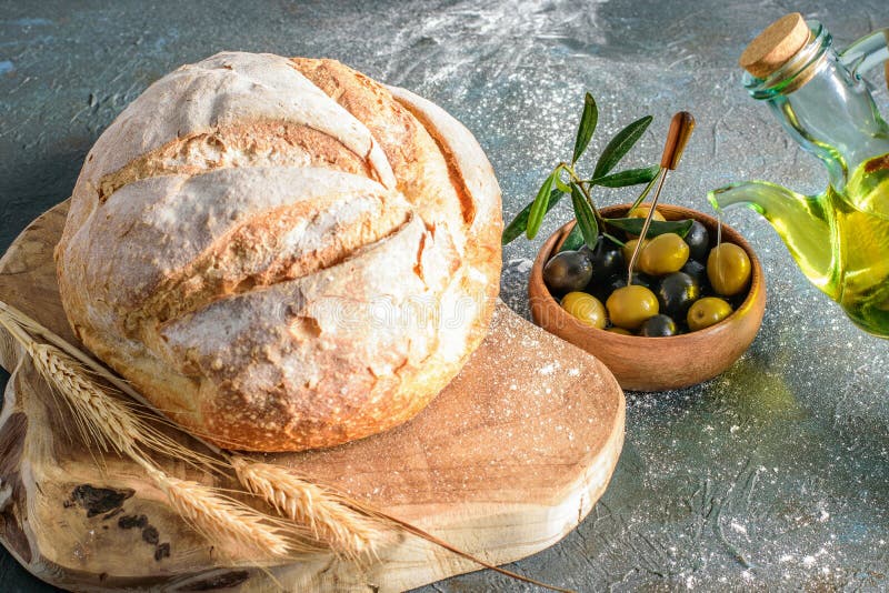 Loaf of white round bread on the cutting board, some wheat ears, olives and olive oil closeup. Healthy homemade white bread