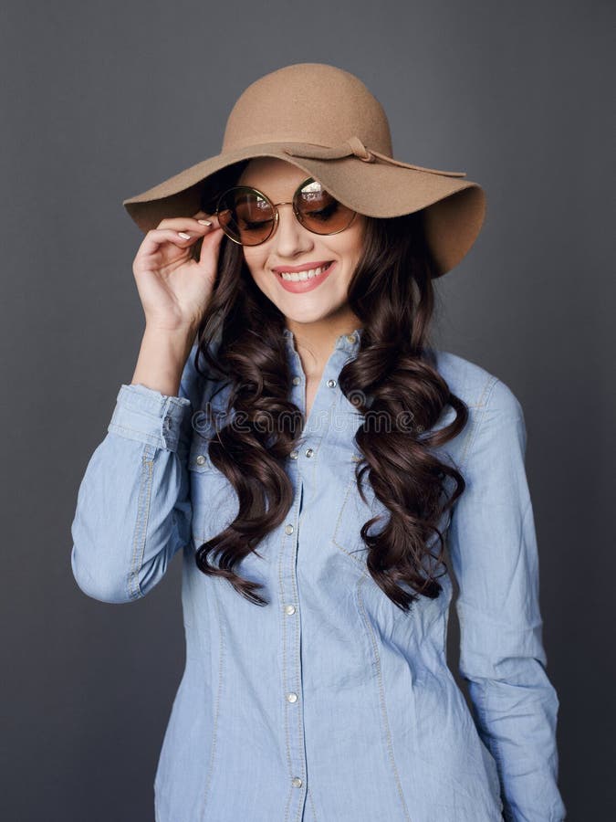 Lockig haired lycklig brunettmodell, elegant iklädd hatt och rund solglasögon, på en grå bakgrund arkivbild Lockig haired lycklig brunettmodell, elegant iklädd hatt och rund solglasögon, på en grå bakgrund arkivbild
