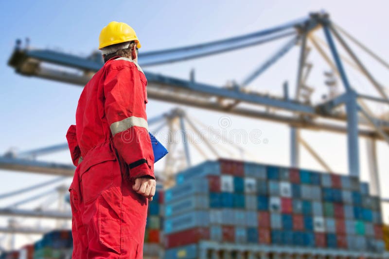 Conceptual image of international logistics, featuring a docker, looking at the unloading of a container ship by huge cranes in the distance. Conceptual image of international logistics, featuring a docker, looking at the unloading of a container ship by huge cranes in the distance
