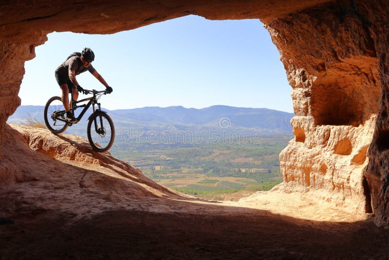 LOKALISIERTER REITER MIT STURZHELM UND RUCKSACK, DER IN EINER MOUNTAINBIKE IN RICHTUNG ZUM EINGANG EINER HÖHLE IM SOMMER ABSTEIGT stockbild