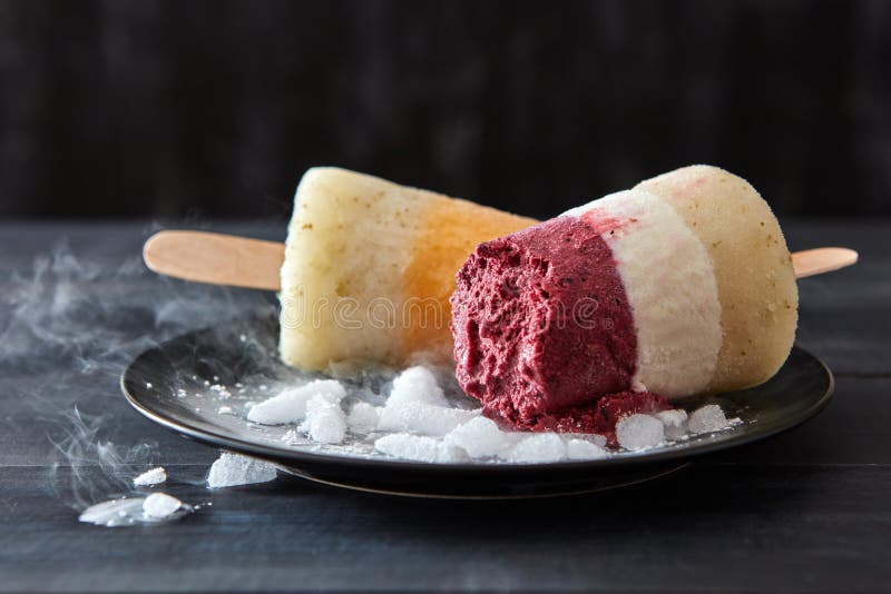 Lolly för glass för fruktkaffe hemlagad på en pinne i en svart platta med skivor av is på en trämörk tabell kallt royaltyfri fotografi Lolly för glass för fruktkaffe hemlagad på en pinne i en svart platta med skivor av is på en trämörk tabell kallt royaltyfri fotografi