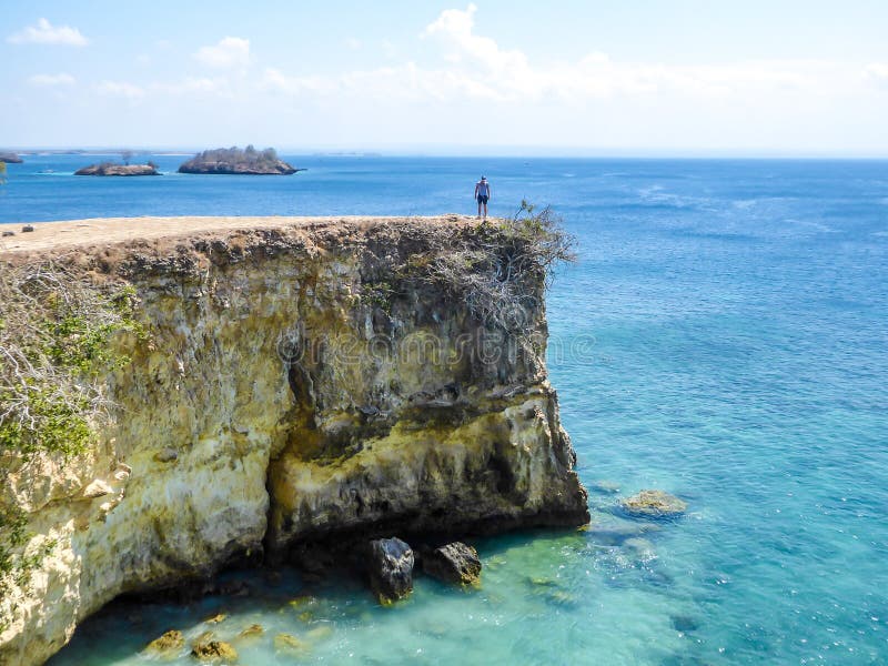 Lombok- мальчик и розовой скала пляжа. Человек в шортах, идущих на краю скалы розового пляжа в Lombook, Индонезии Человек имеет вид Морская вода имеет яркий цвет бирюзового цвета Счастье и авантюрный путешествовать