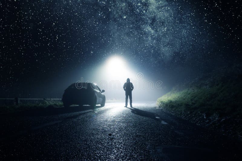 A lone car, parked on the side of the road with a hooded figure, on a spooky country road. With a bright light and stars at night.