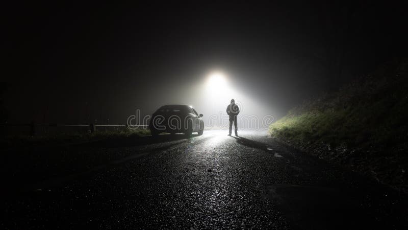 A lone car, parked on the side of the road, underneath a street light, with a hooded figure, on a spooky, scary, rural, country road. On a foggy winters night.